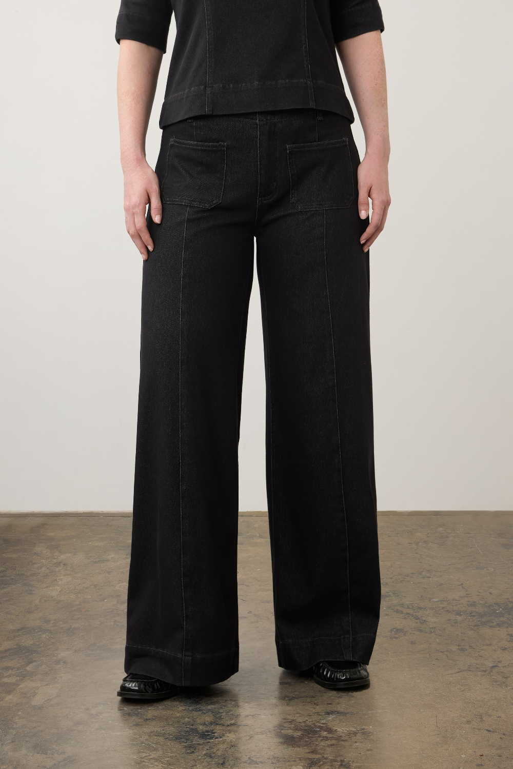 A model from the waist down wearing black wide-leg denim pants, a black top, and black shoes, standing against a neutral studio background.