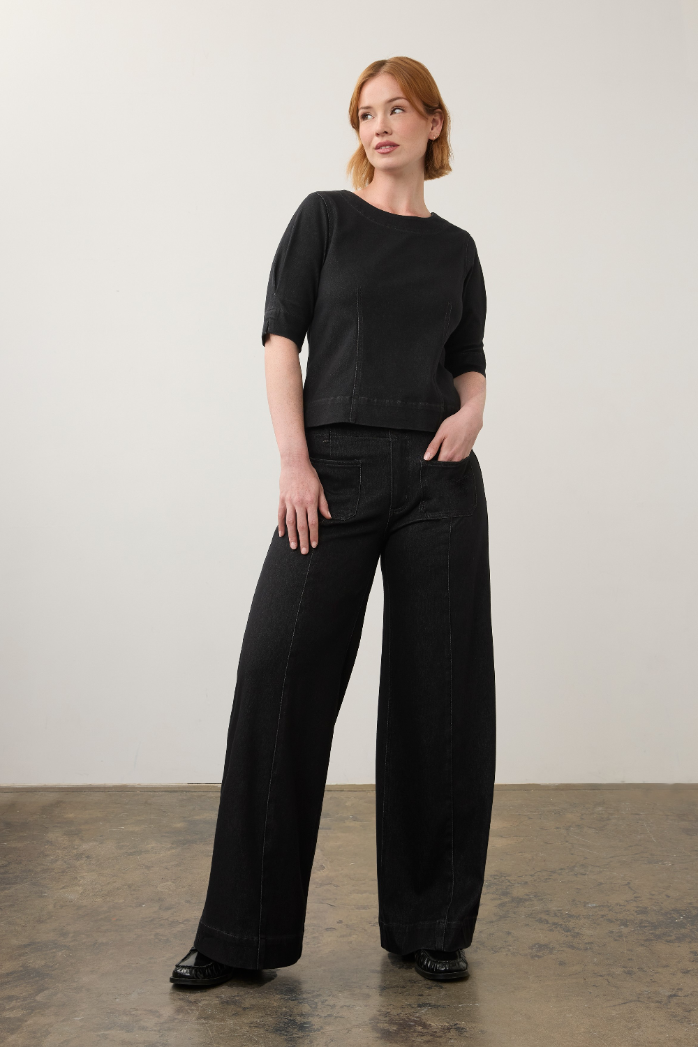 A woman stands in a studio wearing a black short-sleeve top and black wide-leg pants, paired with black shoes, against a plain off-white wall and textured floor.