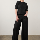 A woman stands in a studio wearing a black short-sleeve top and black wide-leg pants, paired with black shoes, against a plain off-white wall and textured floor.