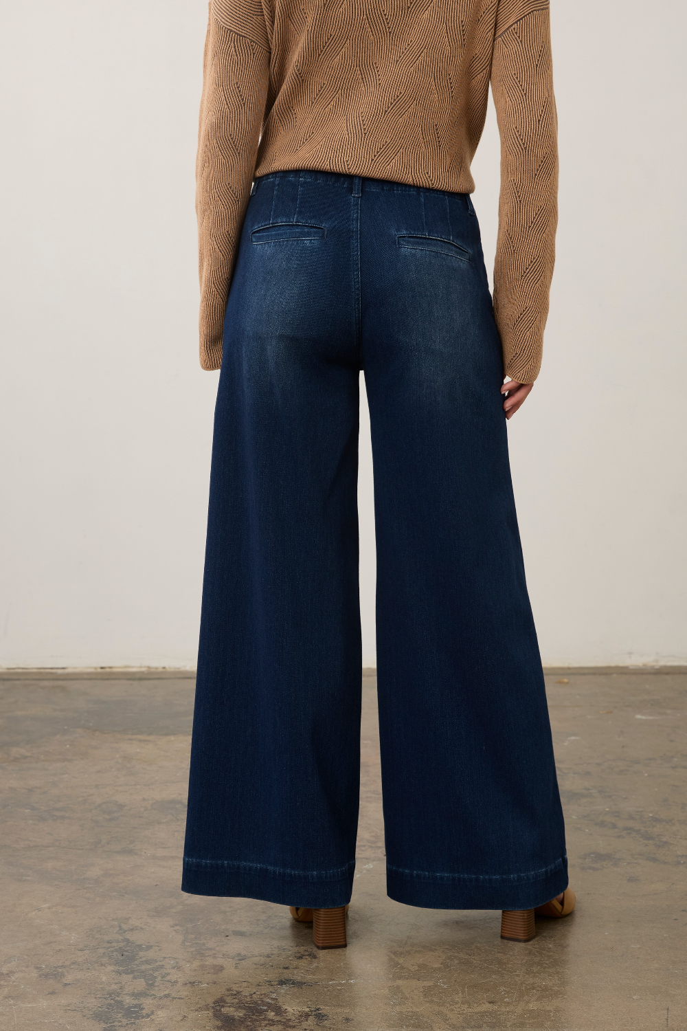 A model viewed from the back wearing dark blue wide-leg denim jeans and a tan cable-knit sweater, standing on a concrete floor against a plain off-white wall.