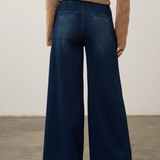 A model viewed from the back wearing dark blue wide-leg denim jeans and a tan cable-knit sweater, standing on a concrete floor against a plain off-white wall.