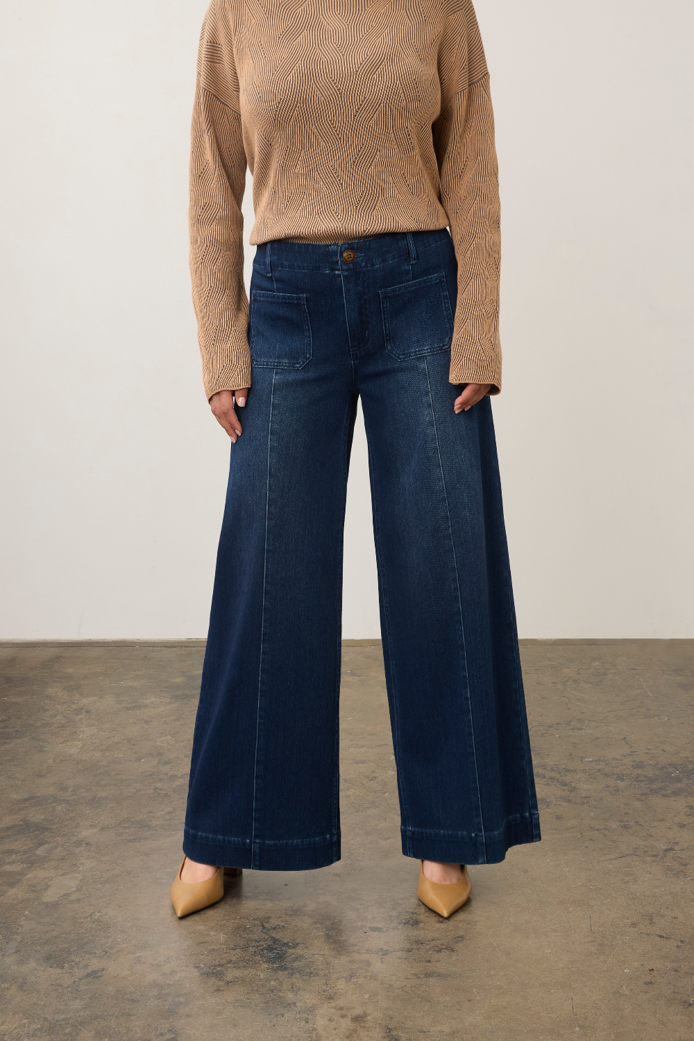 A model wearing dark blue wide-leg denim jeans, a beige textured knit sweater, and beige pointed-toe heels, standing against a neutral studio backdrop.