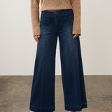 A model wearing dark blue wide-leg denim jeans, a beige textured knit sweater, and beige pointed-toe heels, standing against a neutral studio backdrop.