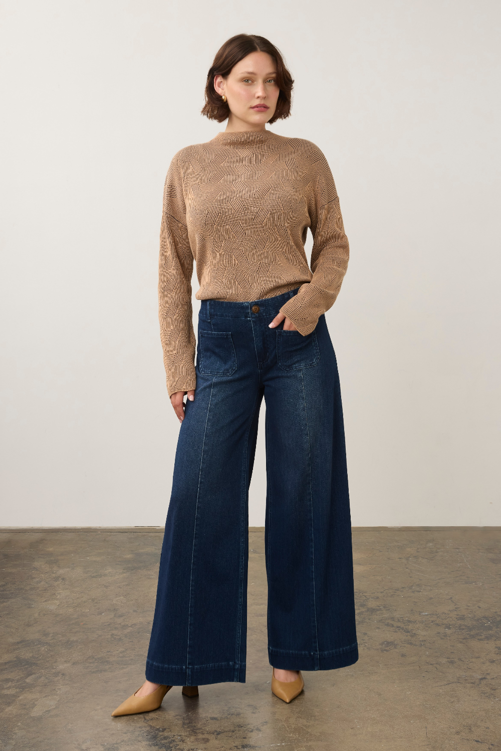 A woman stands in a studio wearing a beige textured mock-neck sweater and dark blue wide-leg jeans, paired with tan pointed-toe heels.