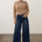 A woman stands in a studio wearing a beige textured mock-neck sweater and dark blue wide-leg jeans, paired with tan pointed-toe heels.