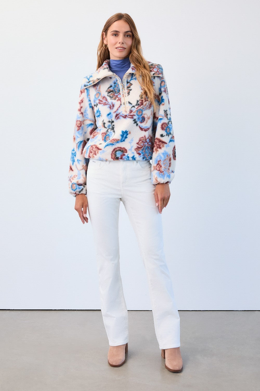 Woman wearing a colorful floral jacket, blue turtleneck, white pants, and pink shoes against a white background