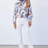 Woman wearing a colorful floral jacket, blue turtleneck, white pants, and pink shoes against a white background