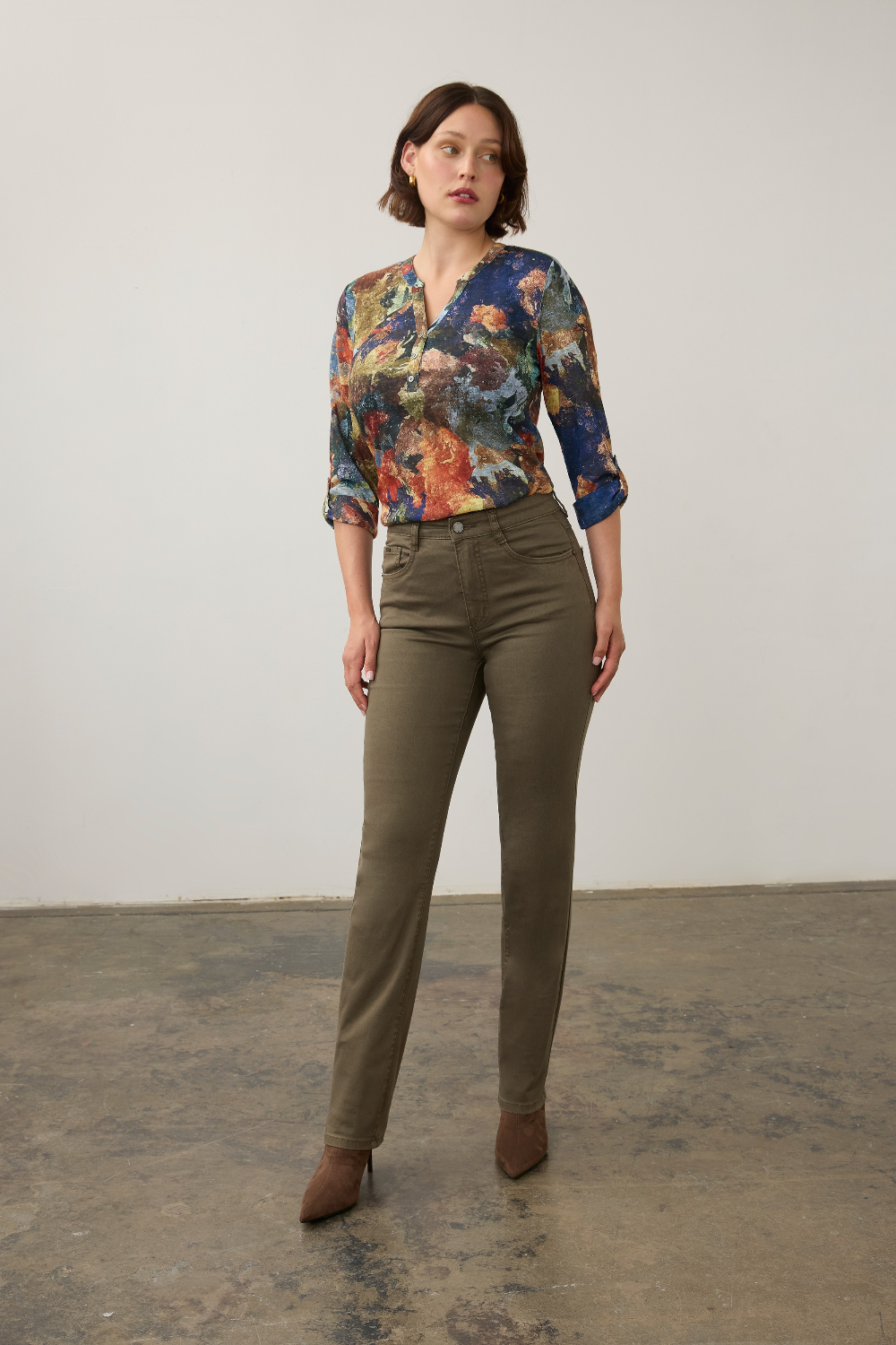 Woman modelling a multi coloured shirt with green jeans  and brown boots photographed against a light background.