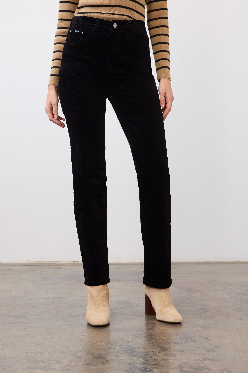 Woman modelling a straight leg black corduroy pant with a tan and black striped sweater and tan boots photographed against a light background.