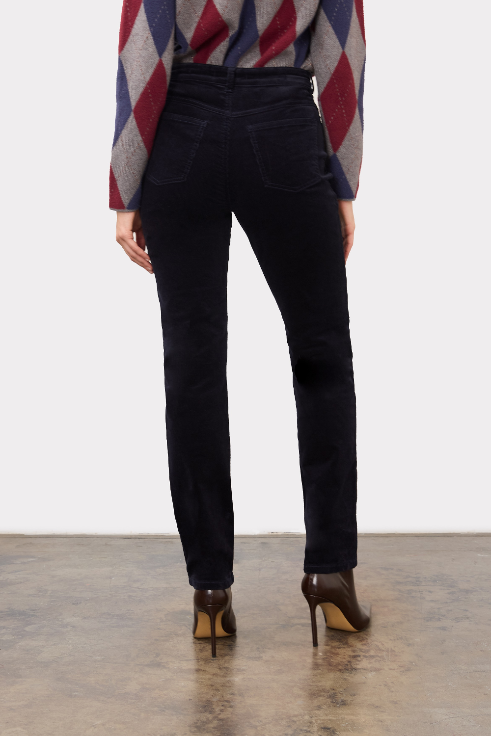 Person, shown from the back, wearing a dark blue straight leg corduroy pant with a grey, blue and burgundy argyle print sweater and brown high heel boots on a neutral background.