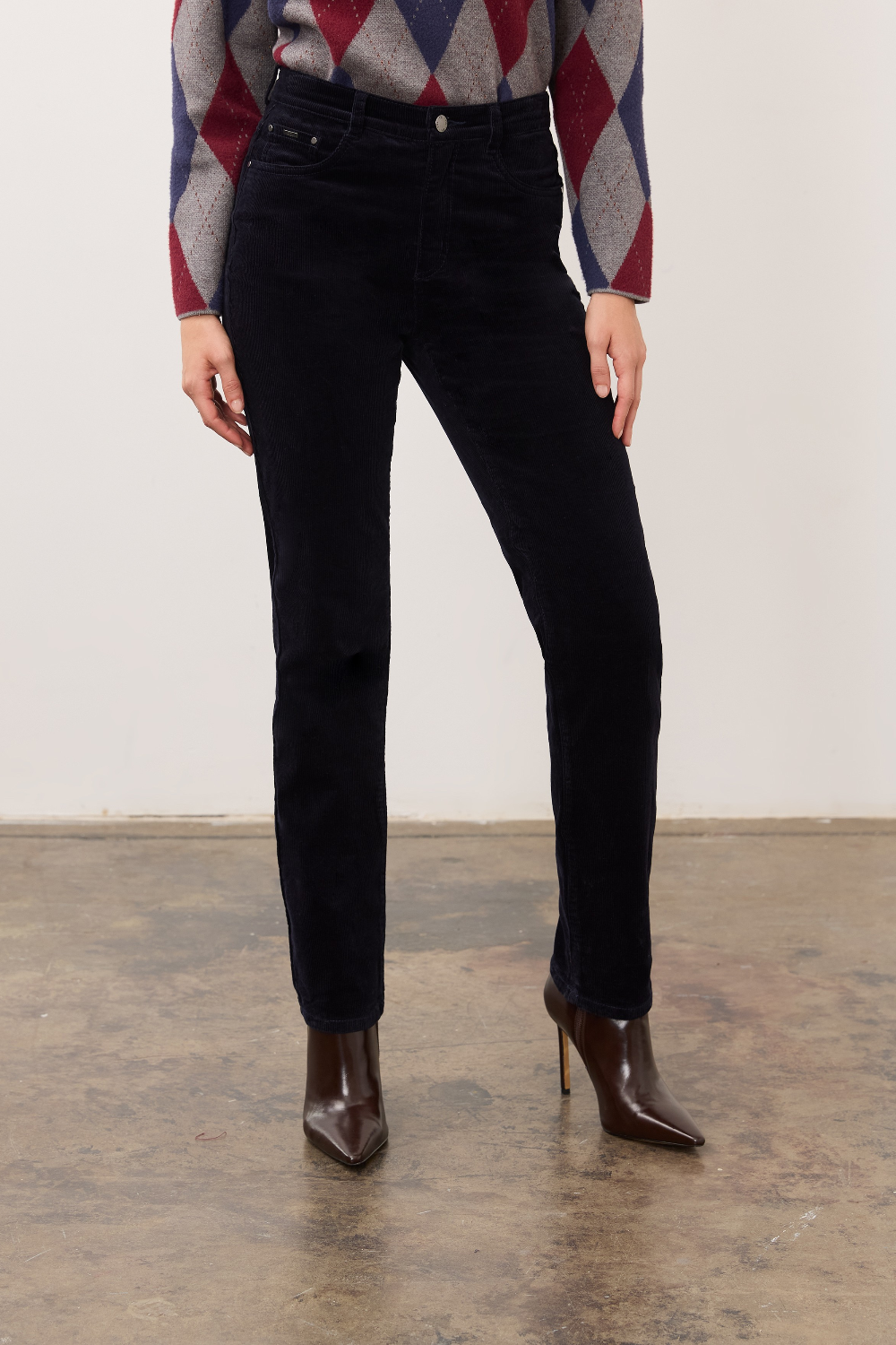 Person wearing a dark blue straight leg corduroy pant with a grey, blue and burgundy argyle print sweater and brown high heel boots on a neutral background.