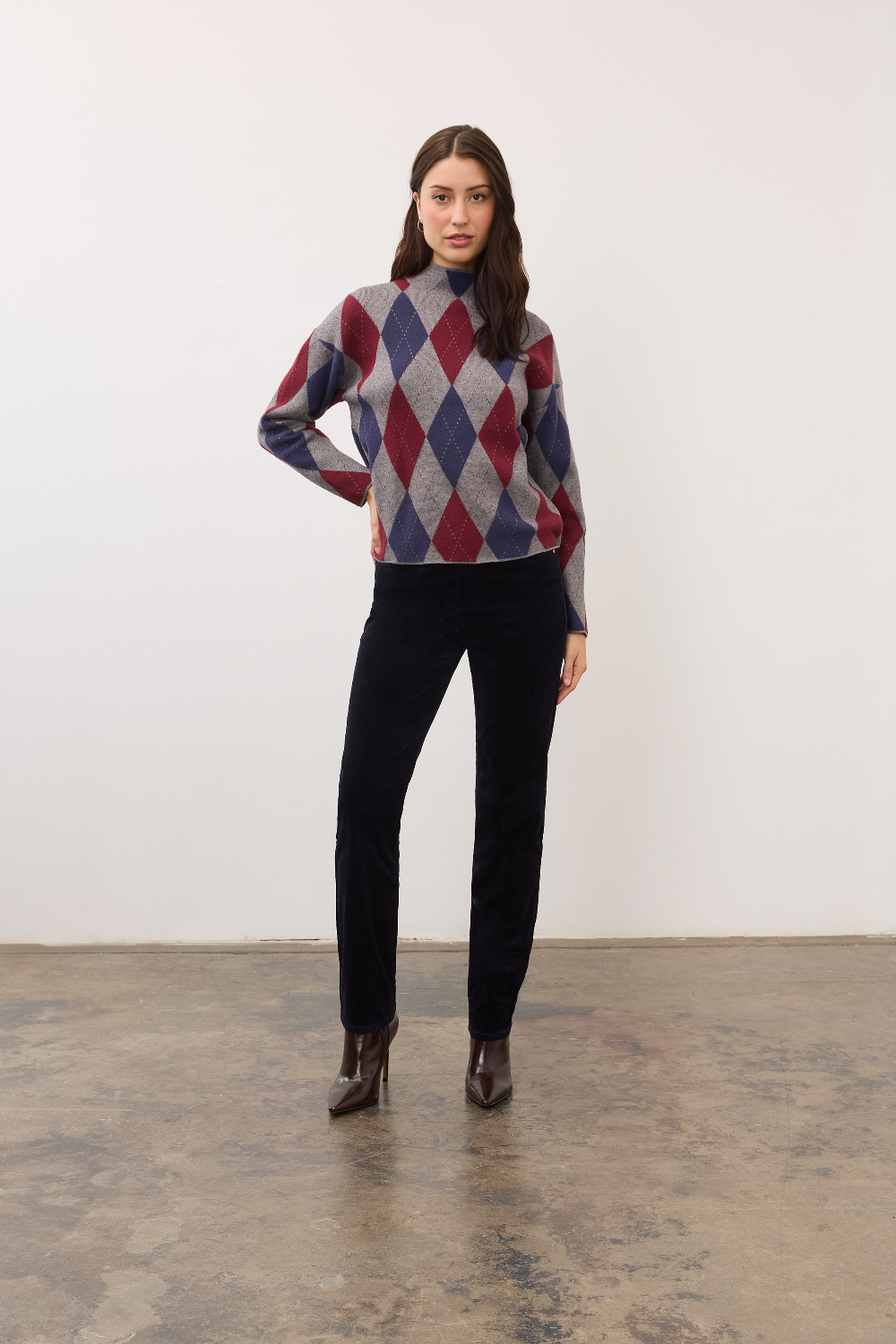 Person wearing a dark blue straight leg corduroy pant with a grey, blue and burgundy argyle print sweater and brown high heel boots on a neutral background.