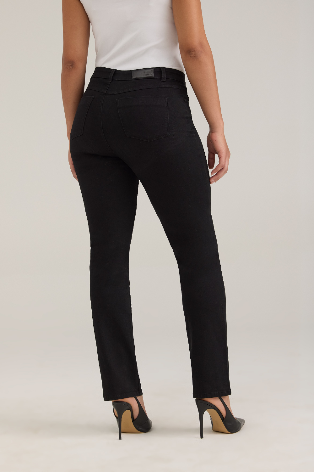 A woman, shown from the back, modeling a pair of black jeans with a white sleeveless top and black high heels, standing against a light gray studio background.