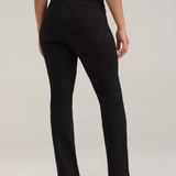 A woman, shown from the back, modeling a pair of black jeans with a white sleeveless top and black high heels, standing against a light gray studio background.