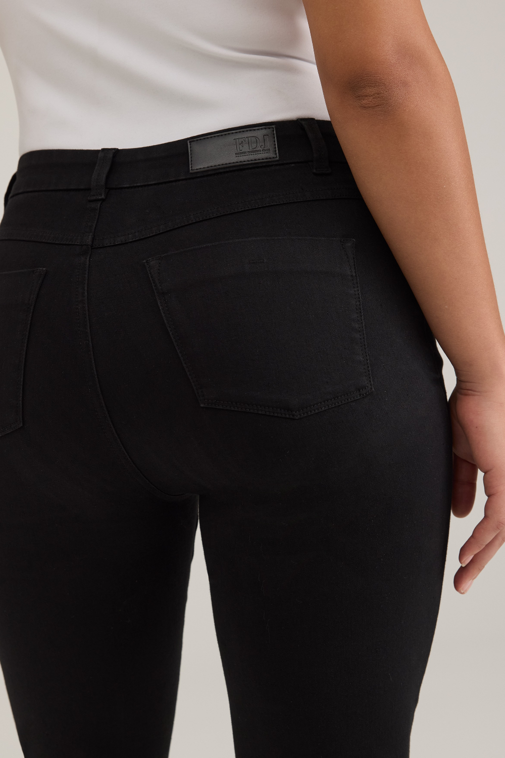 Rear view of a model wearing black high-waisted  jeans and a white top, against a light gray studio background.