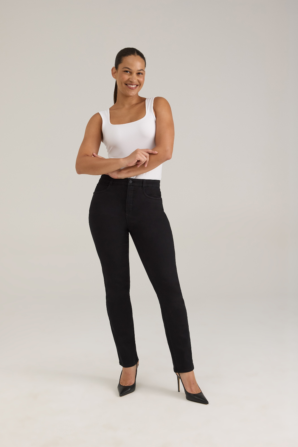 A woman modeling a pair of black jeans with a white sleeveless top and black high heels, standing against a light gray studio background.