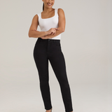 A woman modeling a pair of black jeans with a white sleeveless top and black high heels, standing against a light gray studio background.