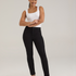 A woman modeling a pair of black jeans with a white sleeveless top and black high heels, standing against a light gray studio background.