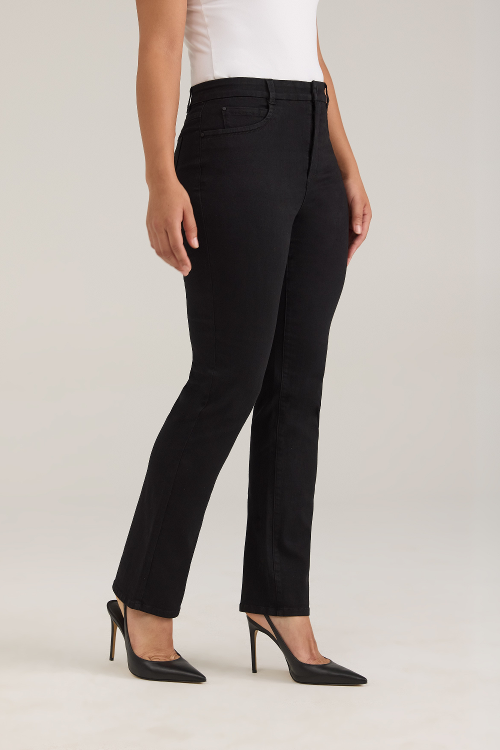 A model wearing black high-waisted straight-leg jeans, styled with a white top and black pointed-toe heels, photographed against a neutral light gray studio background.