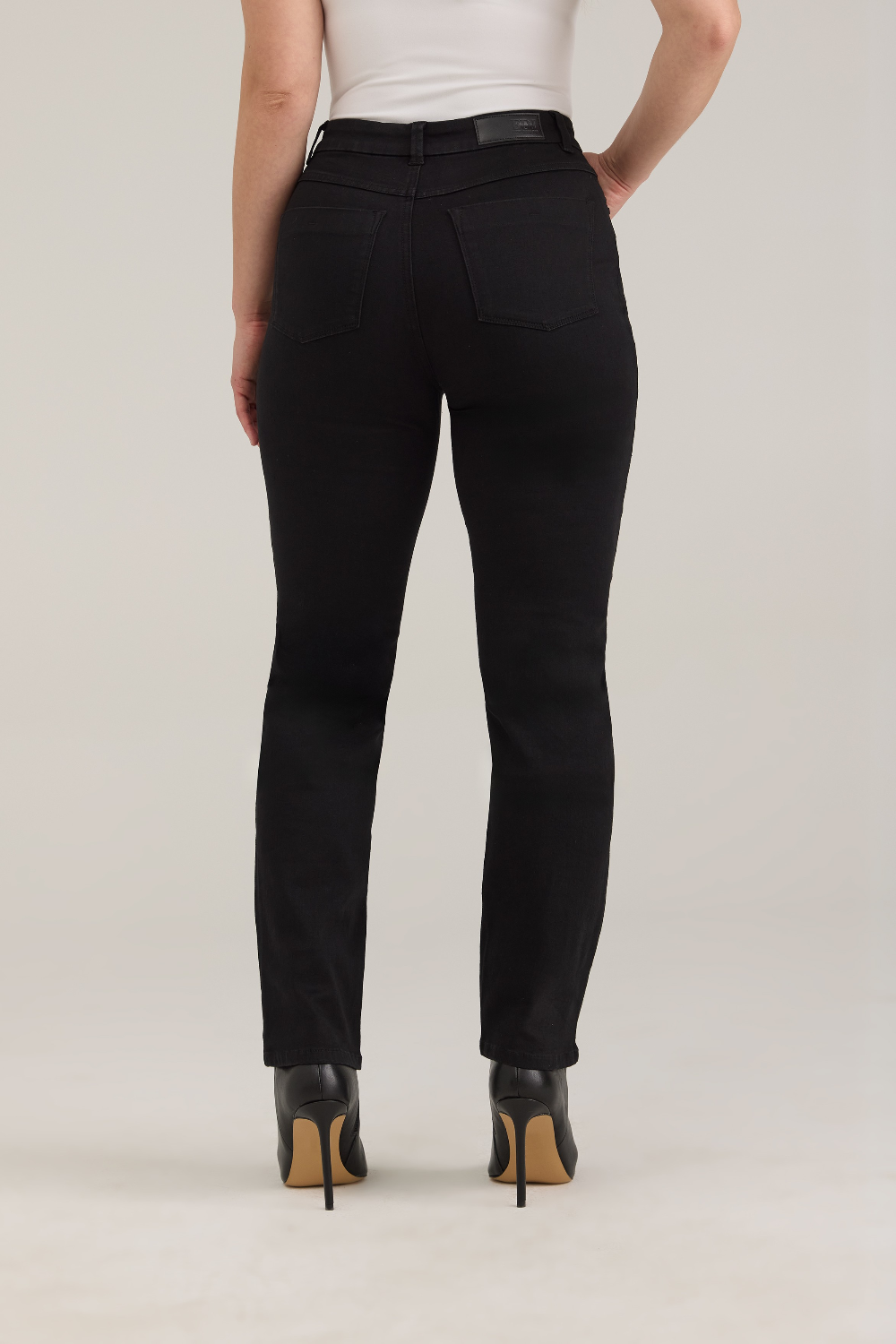 Rear view of a model wearing black high-waisted straight-leg jeans, a white top, and black high-heeled pumps with tan soles, against a light gray studio background.