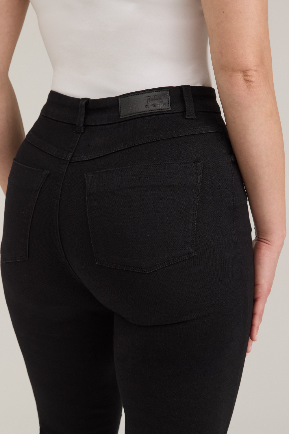 Rear view of a model wearing black high-waisted  jeans and a white top, against a light gray studio background.