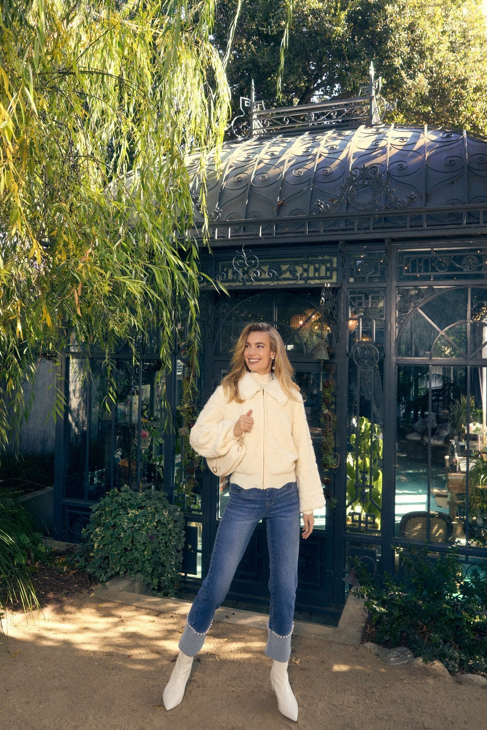 Woman in a cream jacket and blue jeans standing in front of a decorative building with greenery.