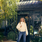 Woman in a cream jacket and blue jeans standing in front of a decorative building with greenery.