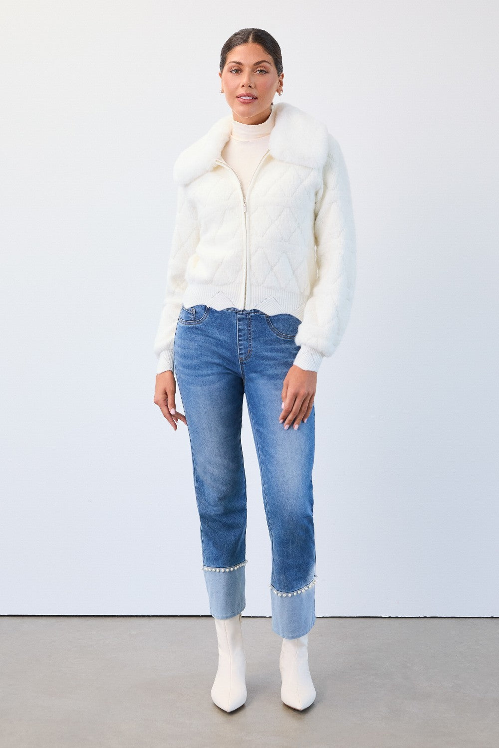 Woman wearing a white puffer vest, cream turtleneck, blue jeans, and white shoes on a plain background