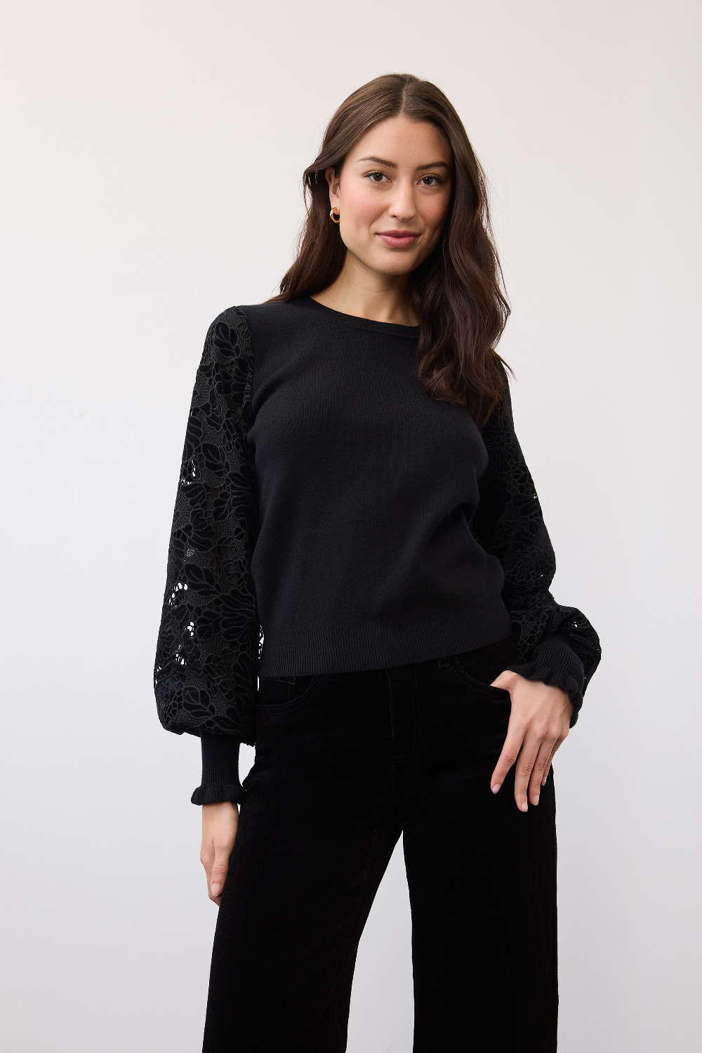 Woman modelling a black crew neck sweater with lace detail on the sleeves with black corduroy pants photographed against a light background.