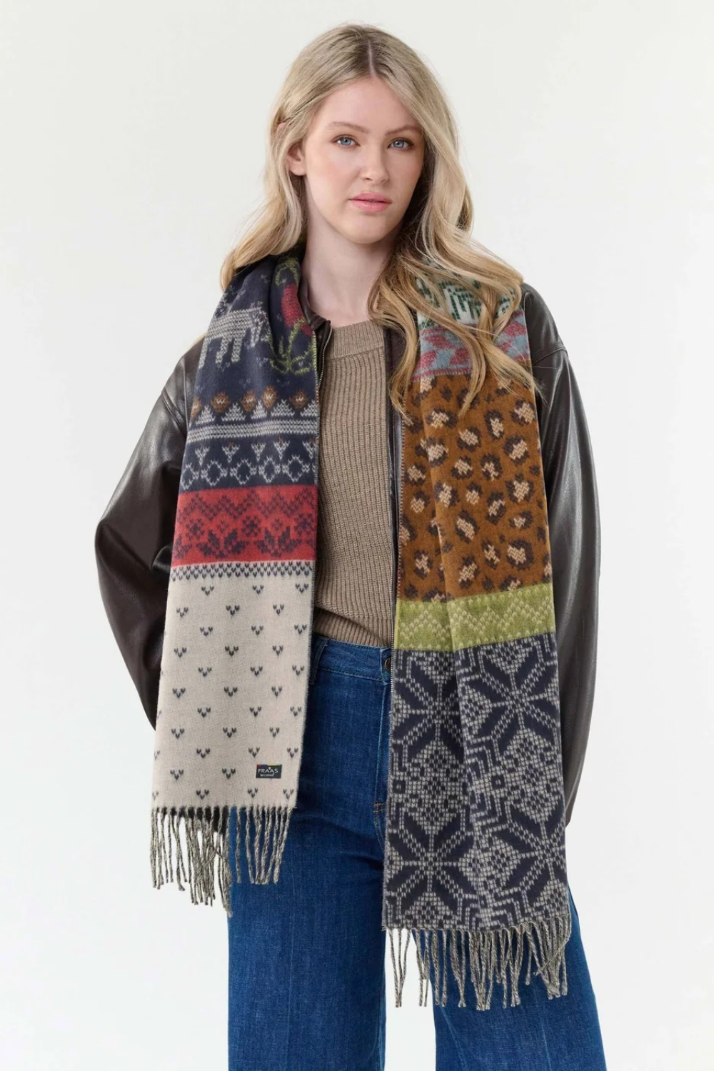 Woman modelling a multi coloured winter themed scarf with a light brown sweater, and blue jeans with a dark brown jacket against a light backdrop.