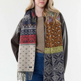 Woman modelling a multi coloured winter themed scarf with a light brown sweater, and blue jeans with a dark brown jacket against a light backdrop.