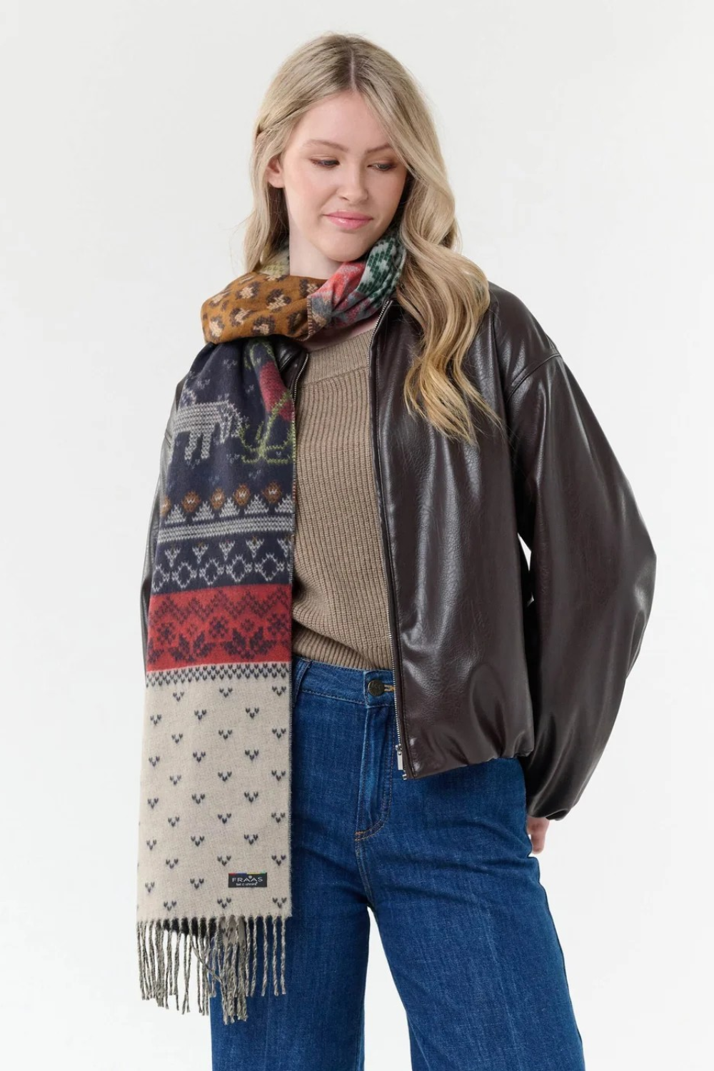Woman modelling a multi coloured winter themed scarf with a light brown sweater, and blue jeans with a dark brown jacket against a light backdrop.