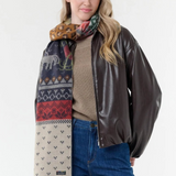 Woman modelling a multi coloured winter themed scarf with a light brown sweater, and blue jeans with a dark brown jacket against a light backdrop.