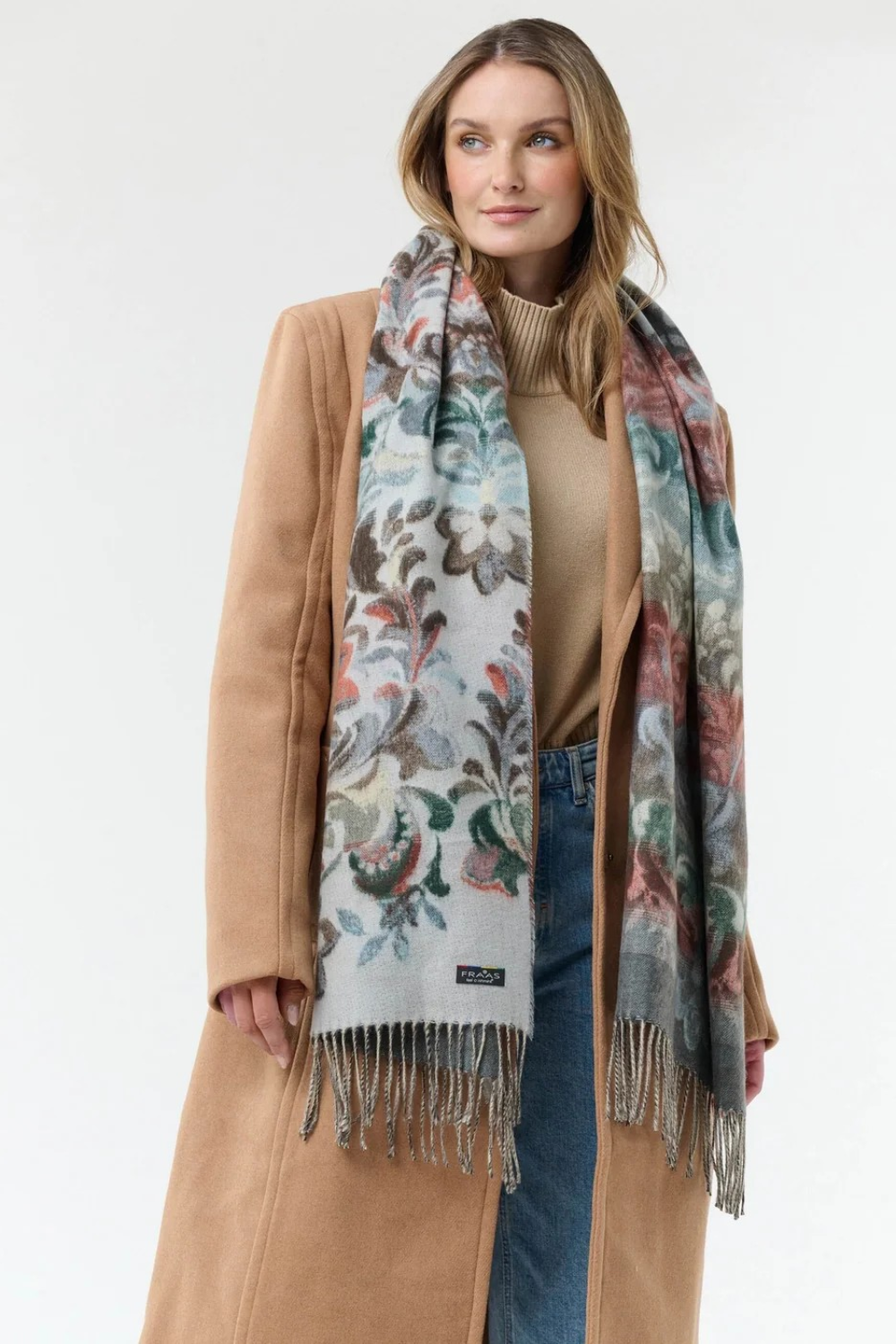 Woman modelling a multi coloured floral scarf, with fringe, with a beige sweater and coat and blue jeans against a light background.