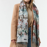 Woman modelling a multi coloured floral scarf, with fringe, with a beige sweater and coat and blue jeans against a light background.