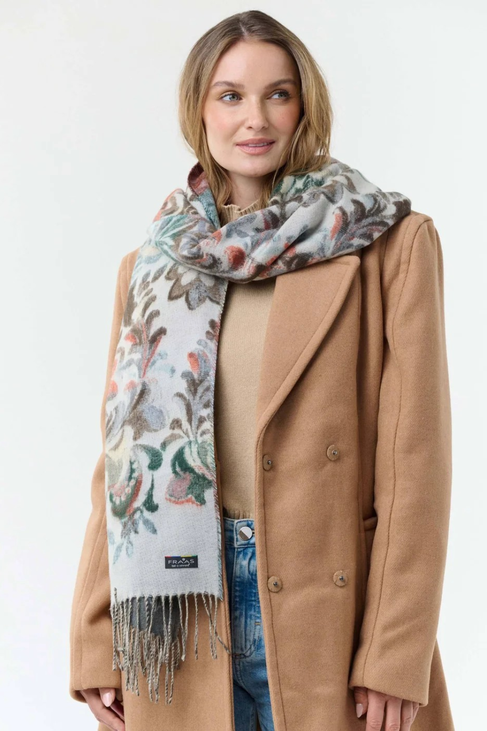 Woman modelling a multi coloured floral scarf, with fringe, with a beige sweater and coat and blue jeans against a light background.