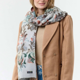 Woman modelling a multi coloured floral scarf, with fringe, with a beige sweater and coat and blue jeans against a light background.