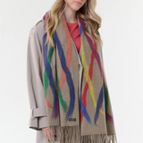 Woman modelling a beige based scarf with multi coloured brushstoke patterns, and fringe, with a grey coat and pink shirt against a light background.
