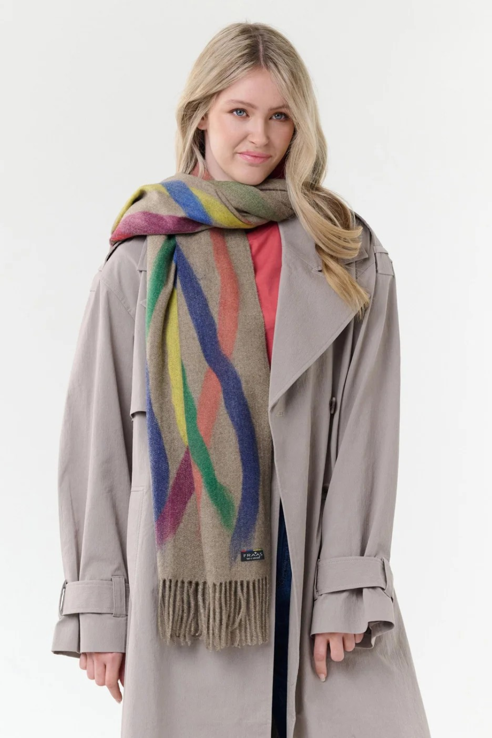 Woman modelling a beige based scarf with multi coloured brushstoke patterns, and fringe, with a grey coat and pink shirt against a light background.