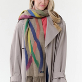 Woman modelling a beige based scarf with multi coloured brushstoke patterns, and fringe, with a grey coat and pink shirt against a light background.