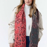 Woman modelling a red and grey paisley scarf, with fringe, with a cream sweater and jacket and blue jeans photographed against a light background.