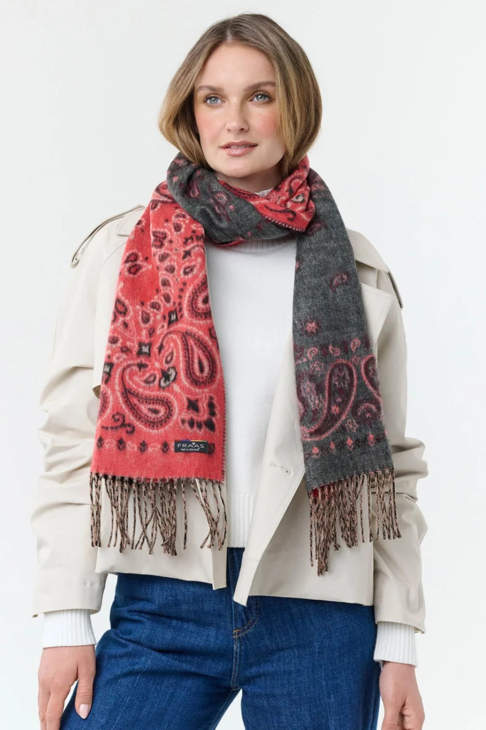 Woman modelling a red and grey paisley scarf, with fringe, with a cream sweater and jacket and blue jeans photographed against a light background.