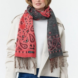 Woman modelling a red and grey paisley scarf, with fringe, with a cream sweater and jacket and blue jeans photographed against a light background.