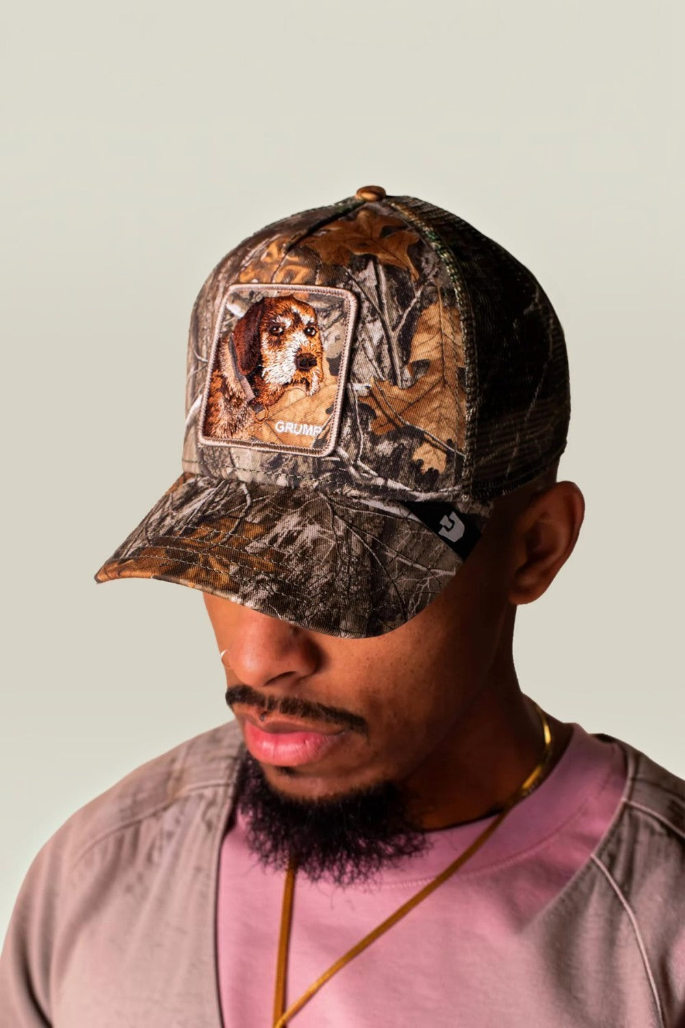 A man wearing a Realtree camouflage baseball cap with a custom dog patch on the front, paired with a pink t-shirt and gold chain against a plain light gray background.