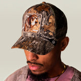 A man wearing a Realtree camouflage baseball cap with a custom dog patch on the front, paired with a pink t-shirt and gold chain against a plain light gray background.