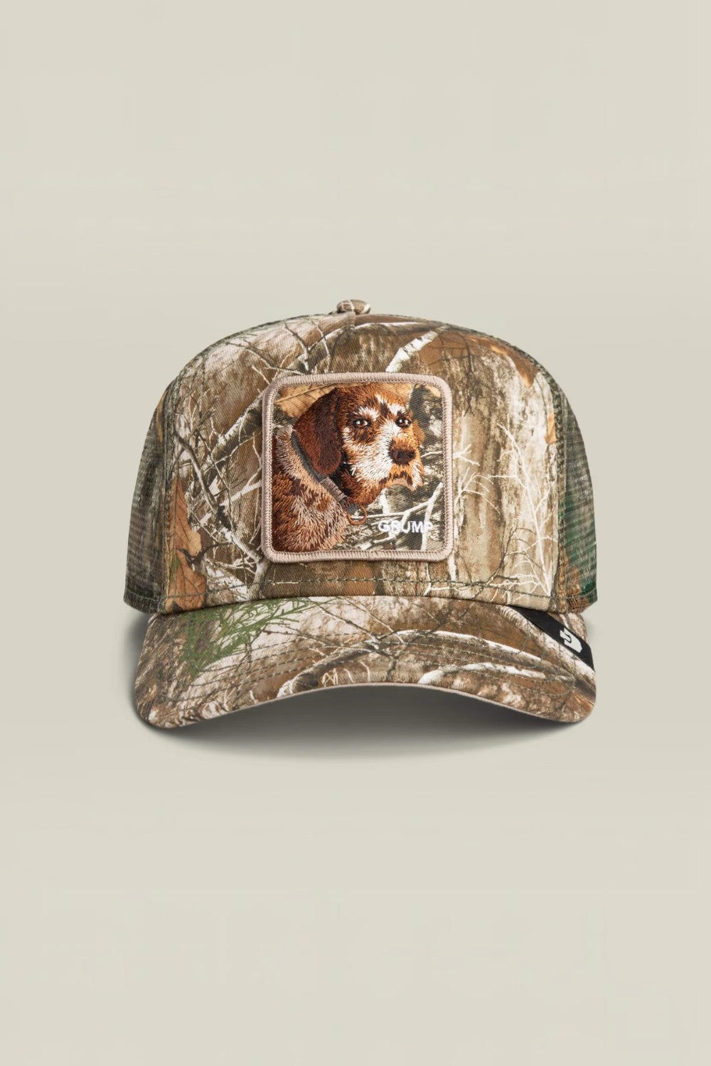 A camouflage trucker hat featuring a square patch on the front with a dog's face, set against a solid light beige background.