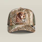 A camouflage trucker hat featuring a square patch on the front with a dog's face, set against a solid light beige background.