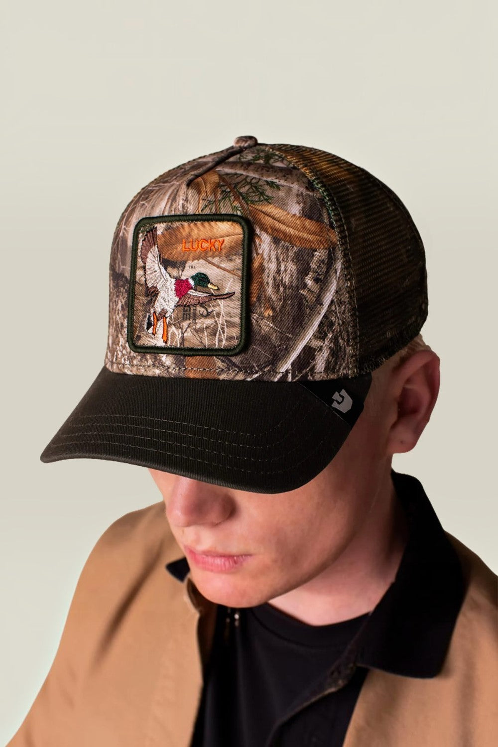 A man wearing a Realtree camouflage trucker hat with a black brim and a front patch featuring a duck hunting scene, paired with a tan jacket over a black shirt, against a plain light gray background.