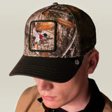 A man wearing a Realtree camouflage trucker hat with a black brim and a front patch featuring a duck hunting scene, paired with a tan jacket over a black shirt, against a plain light gray background.