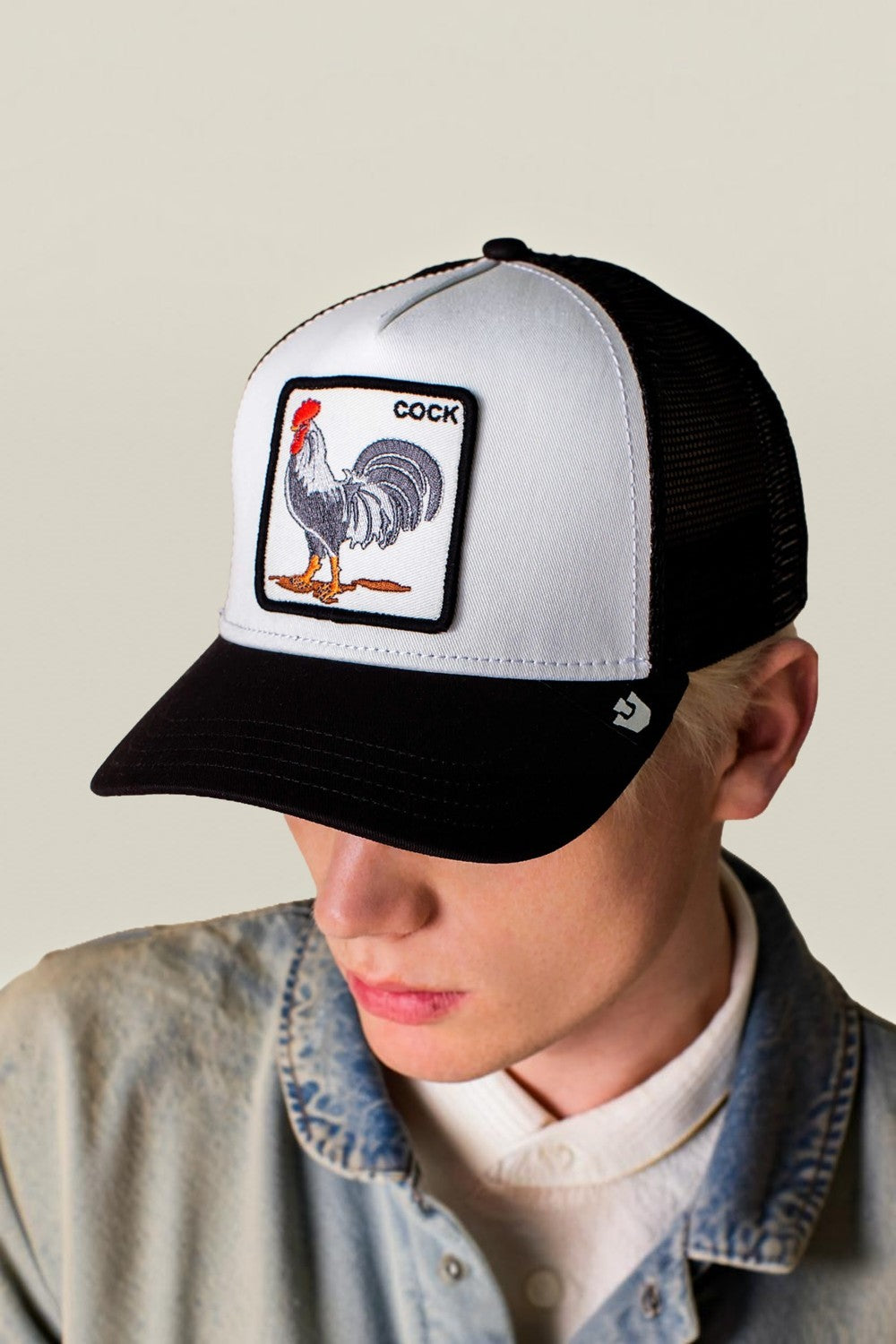A person wearing a black and white trucker hat with a square patch featuring a rooster and the word 'cock'. The photo is cropped from the shoulders up against a plain light beige background.